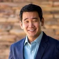 Smiling man in a blue blazer and light blue shirt, standing in front of a brick wall.