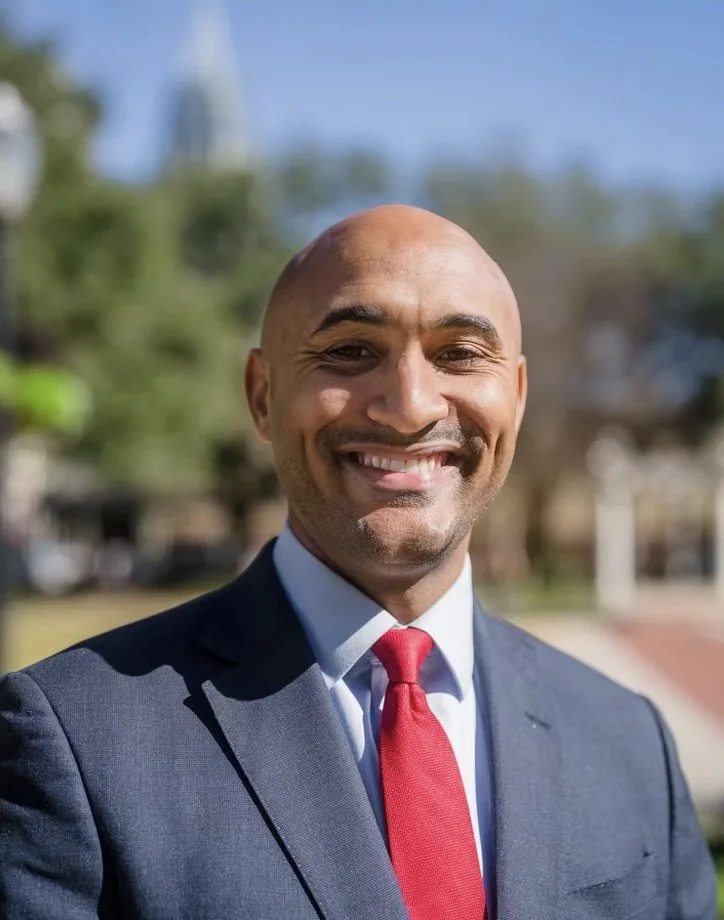 A smiling man in a navy suit, white shirt, and red tie standing outdoors with blurred trees and buildings in the background.