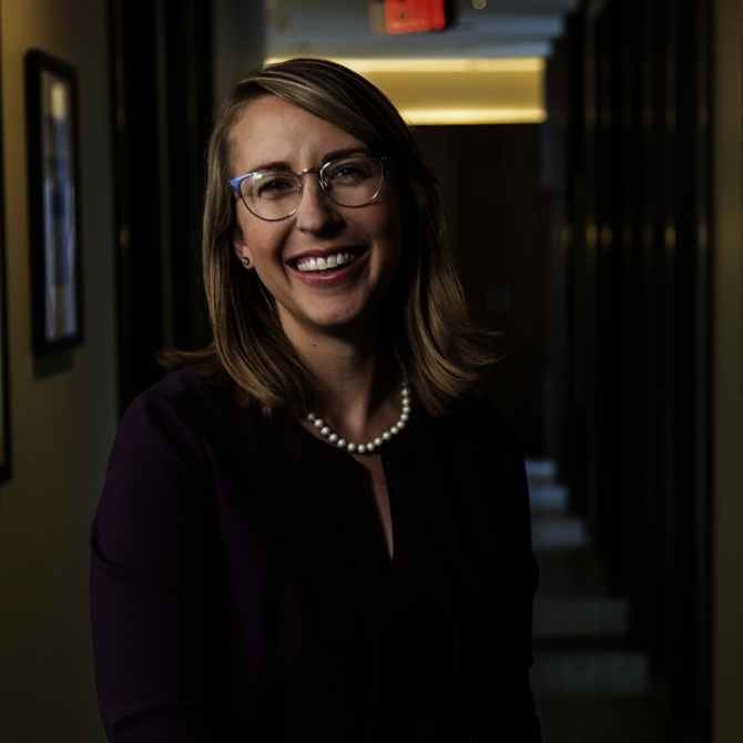 Smiling woman with shoulder-length light brown hair, wearing glasses, a dark purple blazer, and a pearl necklace, standing in a hallway.