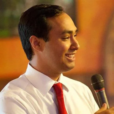 A man smiling while holding a microphone, dressed in a white shirt and red tie, in an indoor setting.