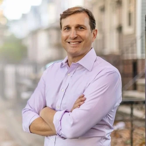 A man with light brown hair and a friendly smile, wearing a light purple button-up shirt, standing outdoors on a city street with buildings and a blurred background.