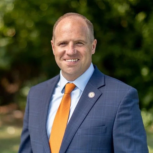 A smiling man in a blue suit, white shirt, and orange tie standing outdoors with green trees in the background.