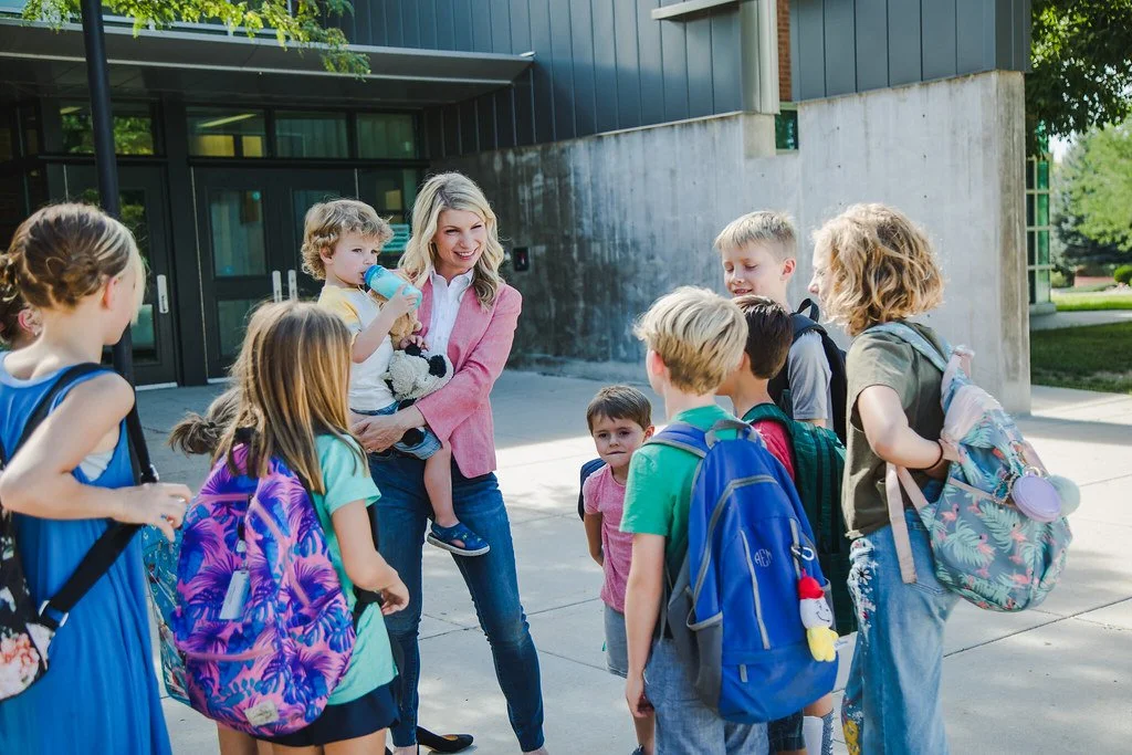 A woman with blonde hair, wearing a pink blazer, stands outside a school building talking to a group of young children with backpacks.