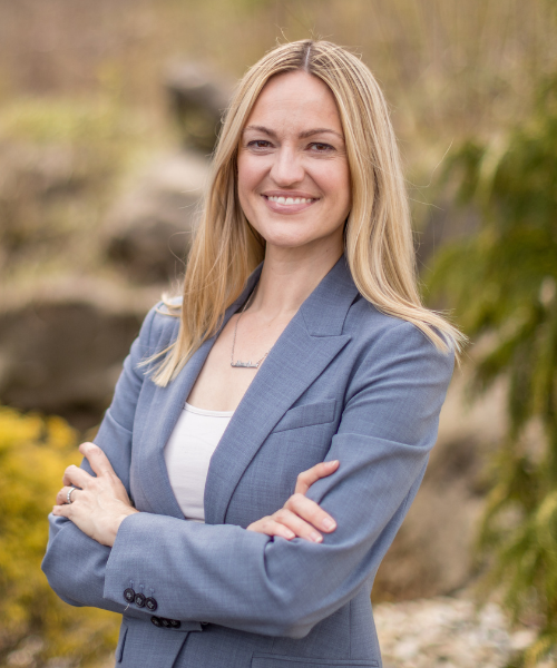 A woman with long blonde hair wearing a blue blazer and white top, smiling with arms crossed outdoors in a natural setting.