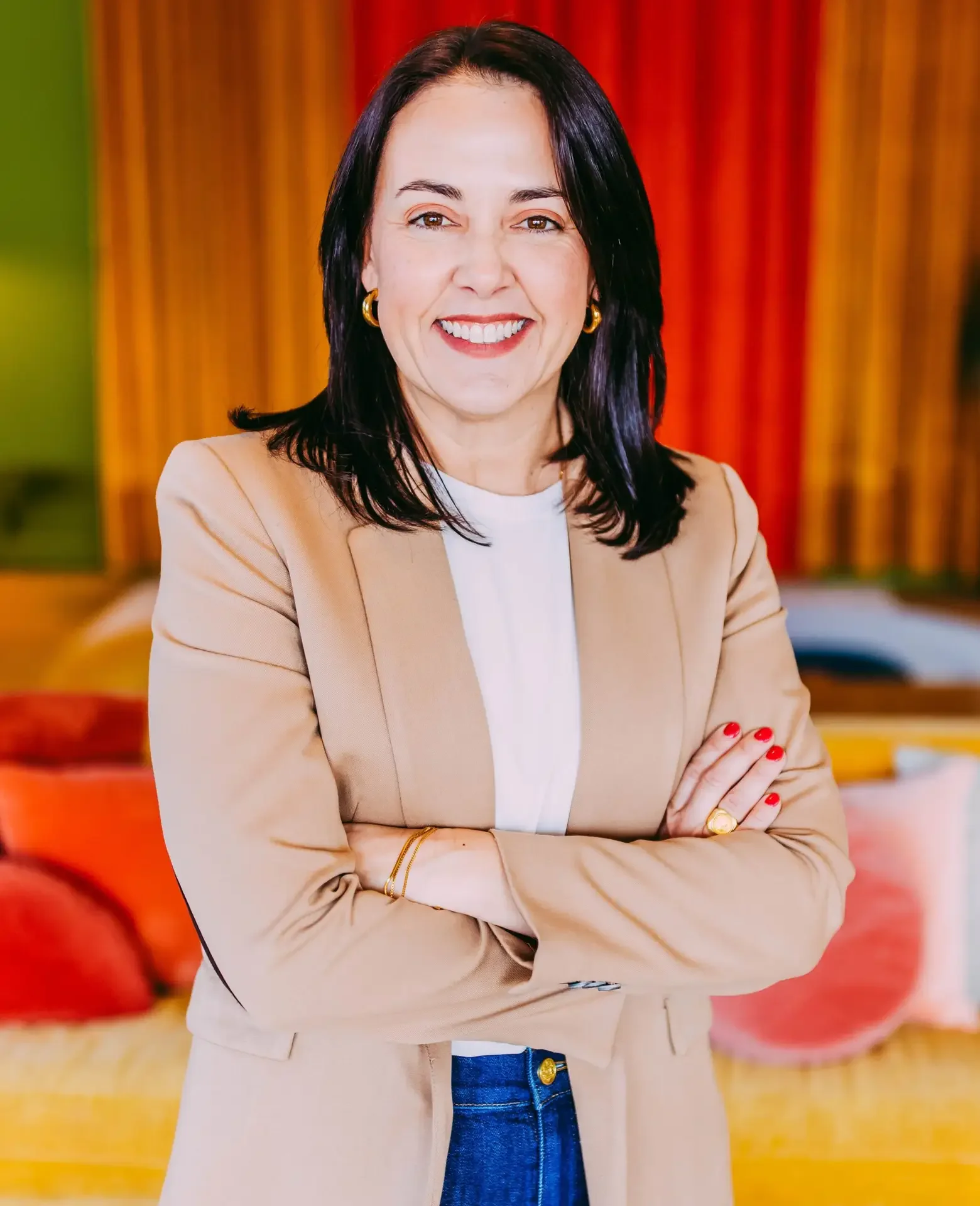 Woman with black hair smiling, wearing a beige blazer, white top, blue jeans, gold jewelry, standing with arms crossed. Colorful background with red and orange curtains.
