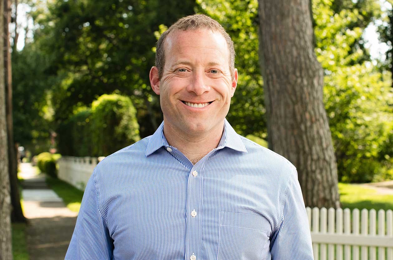 A man smiling outdoors in front of trees and a white picket fence, wearing a blue checkered button-up shirt.