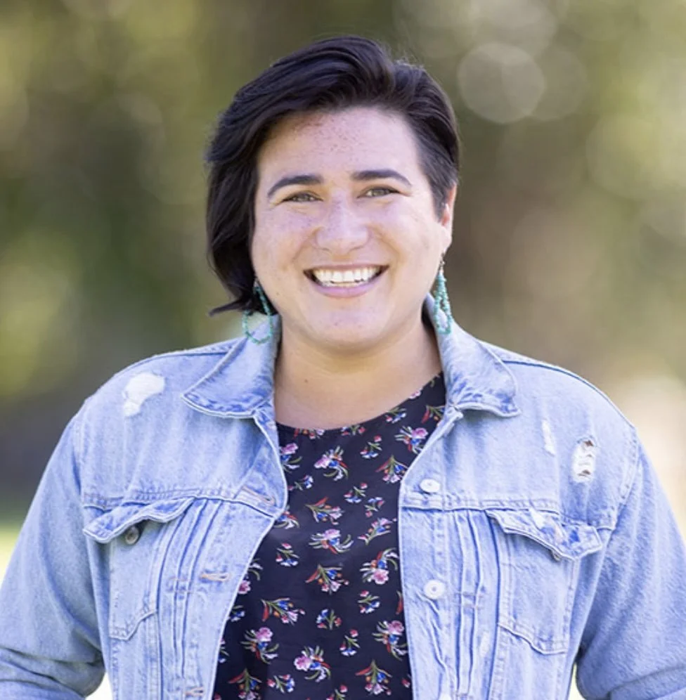 A young woman with short dark hair smiling outdoors, wearing a distressed denim jacket over a black floral top, with turquoise earrings, in a natural setting with blurred greenery in the background.