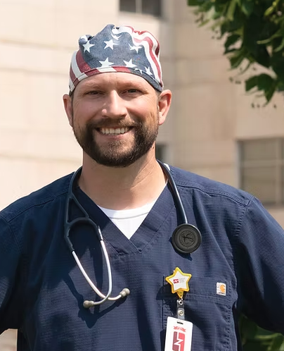 A smiling male healthcare professional wearing a navy scrubs, a stethoscope, an American flag bandana, and a nurse badge, outdoors with trees and a building in the background.