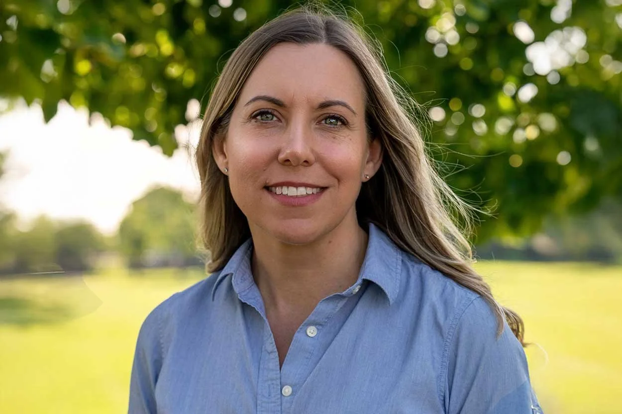 A woman with long brown hair wearing a light blue button-up shirt standing outdoors in a park with green trees and grass in the background, smiling.