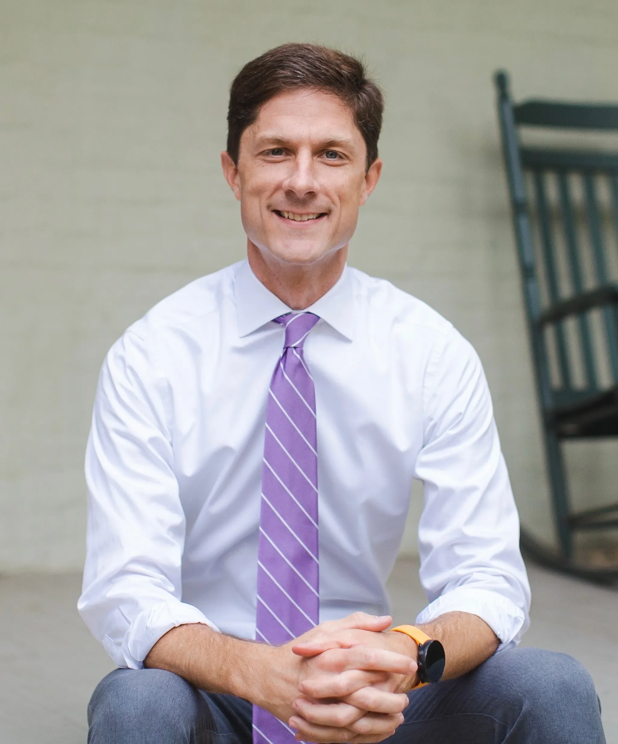 A man with short brown hair, wearing a white dress shirt, purple striped tie, and a black watch, sitting with his hands clasped, smiling in front of a greenish wall and a black chair.