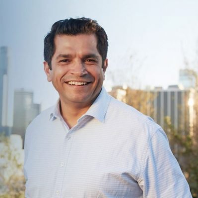 A smiling man in a white dress shirt standing outdoors with a city skyline in the background.