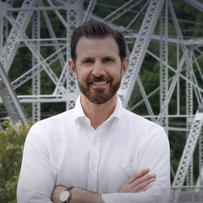 A man with a beard and short dark hair smiling in front of a steel bridge structure with trees in the background