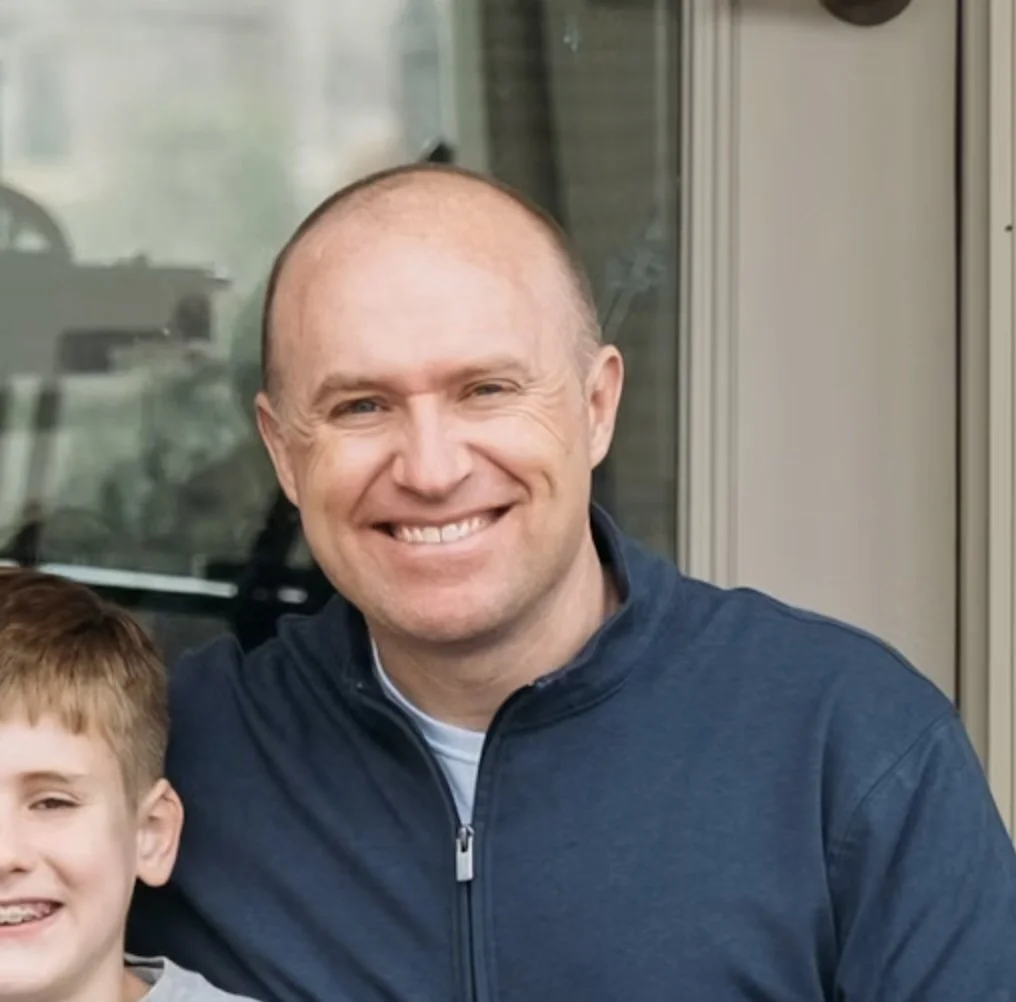 A smiling man with a shaved head wearing a navy blue zip-up jacket, standing next to a young boy with short red hair and a big smile, in front of a glass door or window.