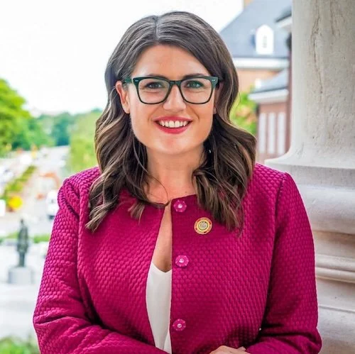 A woman with long dark hair, glasses, and a pink textured blazer smiling outdoors with a blurred cityscape background.