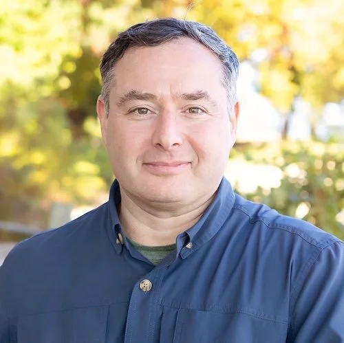 A man with short dark hair and fair skin, wearing a blue button-up shirt, standing outdoors with trees and sunlight in the background.