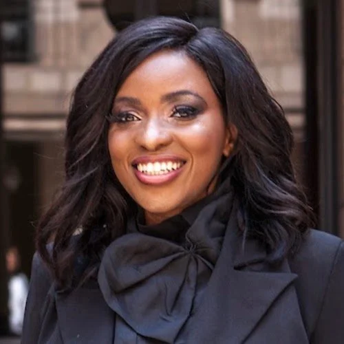 A woman with dark hair and makeup smiling, wearing a black jacket with a scarf, standing outdoors in front of a building.