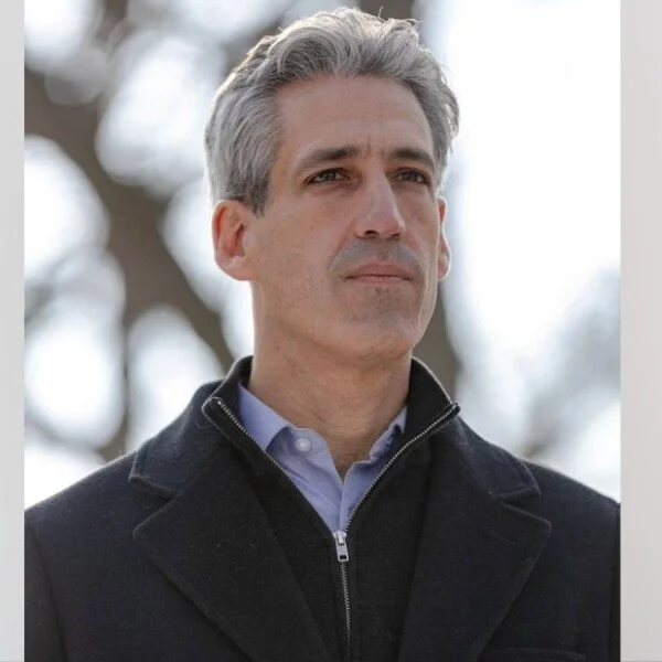 A middle-aged man with gray hair standing outdoors, wearing a dark coat over a light blue collared shirt, with a serious expression, against a background of blurred trees.