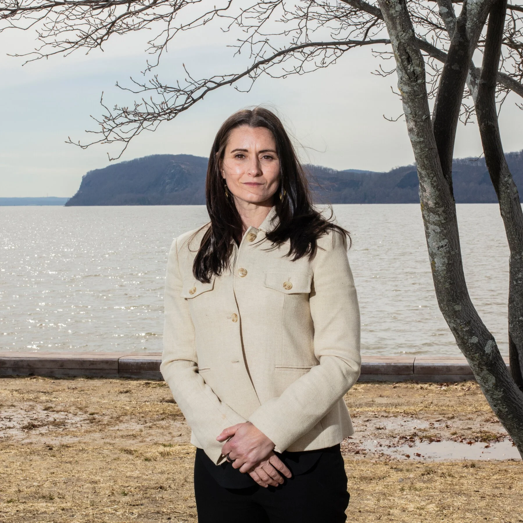 A woman with dark hair standing outdoors near a tree by the water with mountains in the background, wearing a light-colored coat.