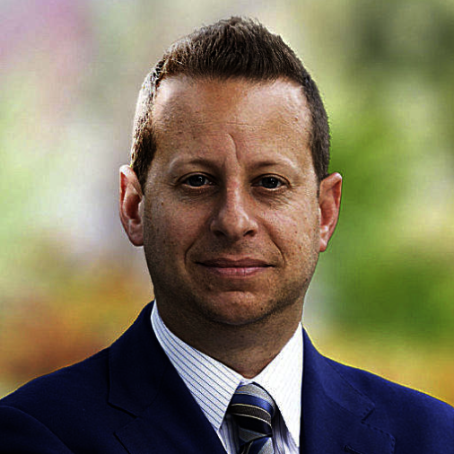 A man with short brown hair in a business suit standing outdoors with blurred colorful background.
