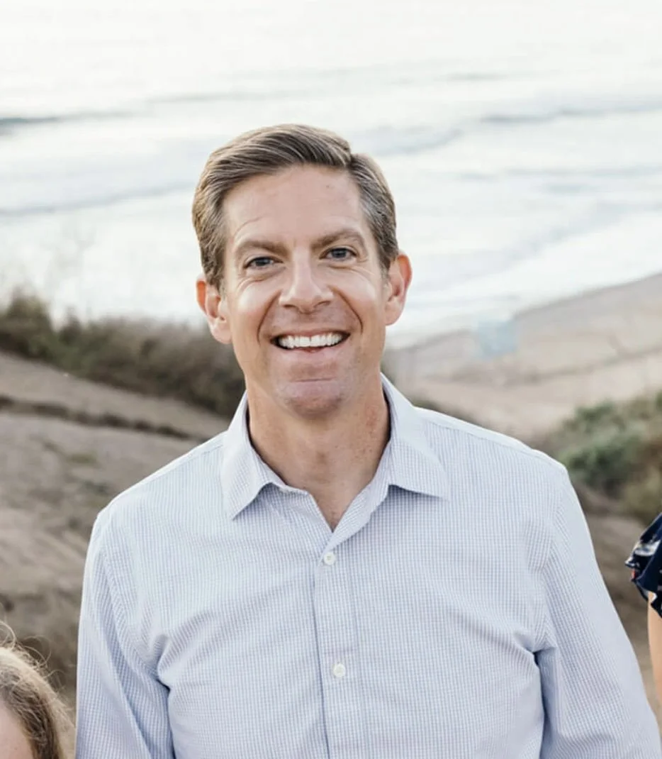 A smiling man in a light blue button-up shirt, standing outdoors near a body of water, possibly at a beach or lakeside.