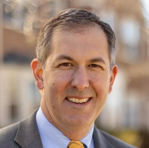 A smiling man with gray hair wearing a gray suit, light blue shirt, and yellow tie, outdoors in a residential neighborhood.