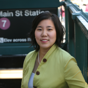 Woman with shoulder-length dark hair wearing a light green jacket, standing near a subway entrance in an outdoor urban setting.