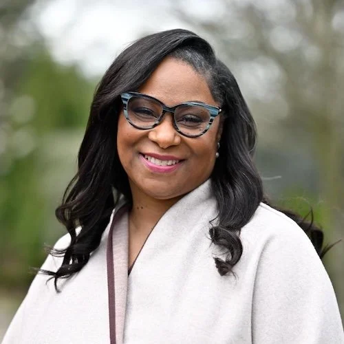 A woman with black wavy hair wearing glasses and a light-colored coat, smiling outdoors.