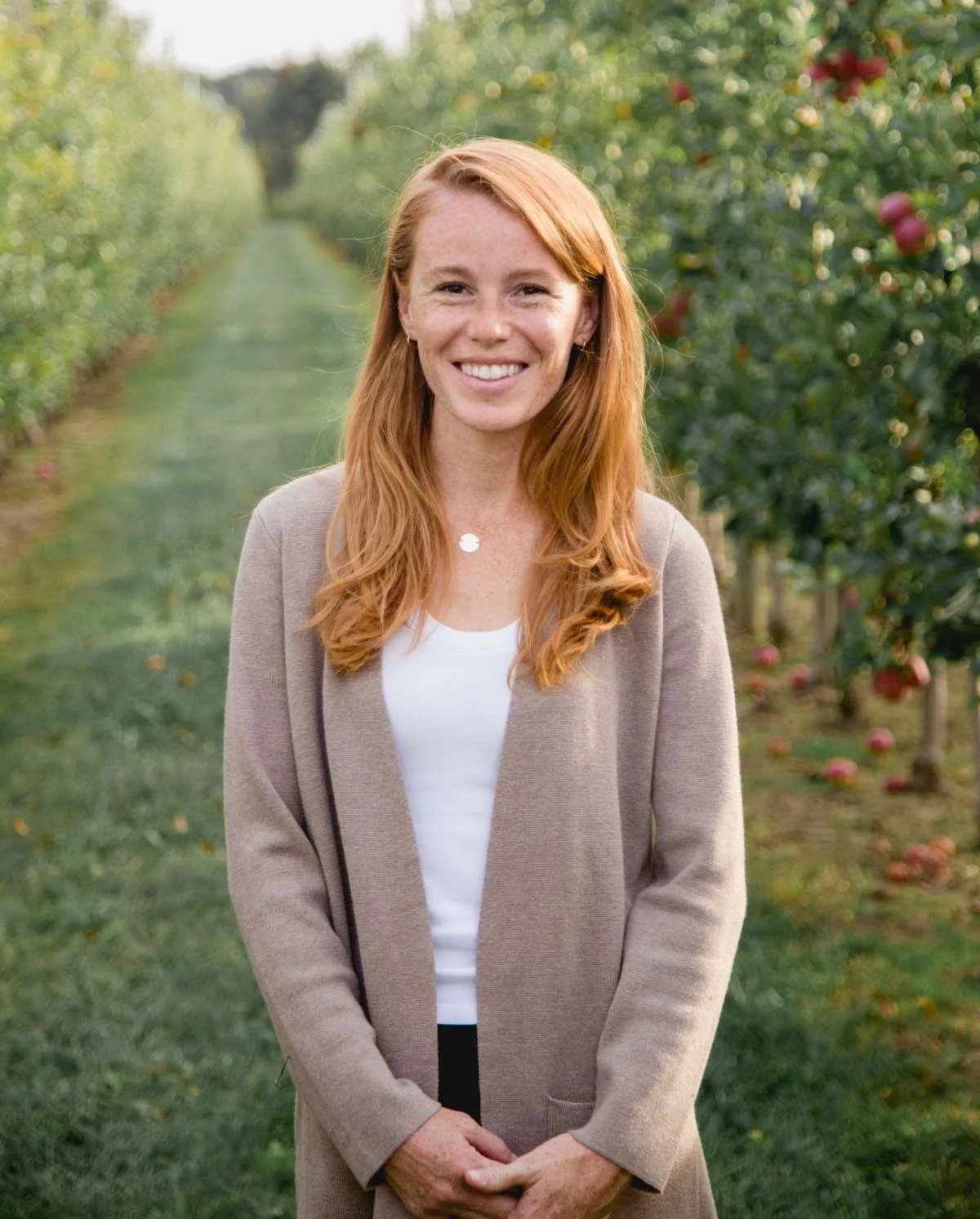 A woman with long red hair smiling outdoors in an apple orchard, wearing a beige blazer over a white top.