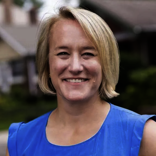 A woman with short blonde hair smiling outdoors, wearing a blue sleeveless top with a blurred house and greenery in the background.