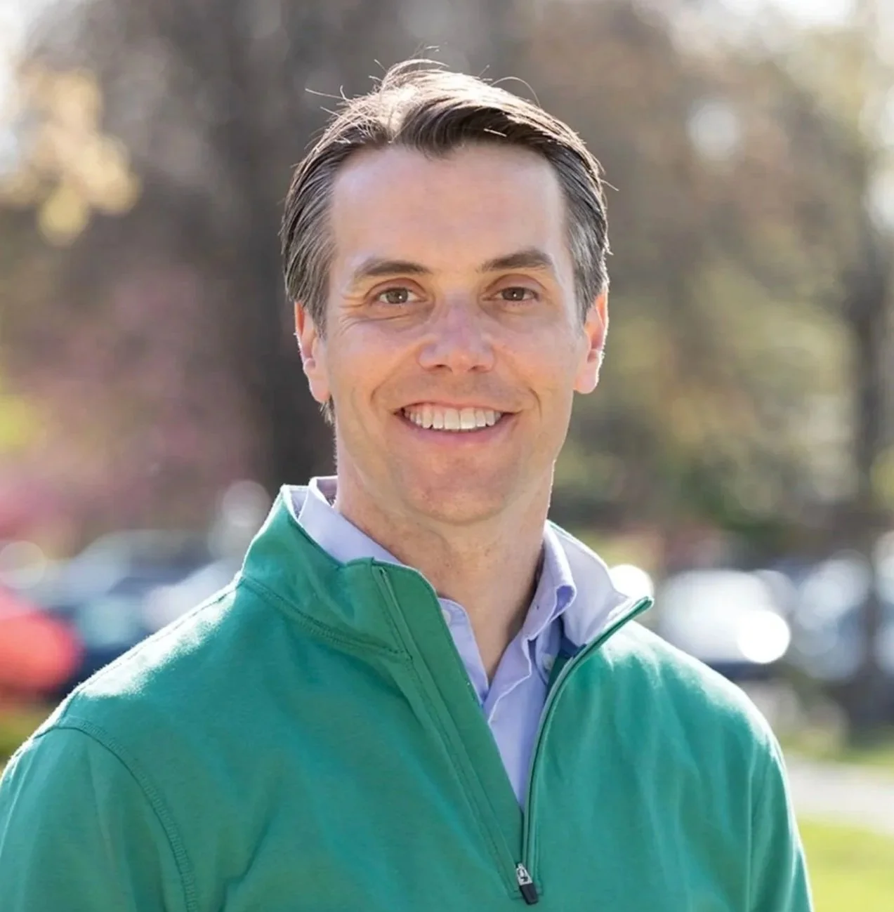 A smiling man with short brown hair, wearing a green zip-up jacket over a collared shirt, outdoors on a sunny day.