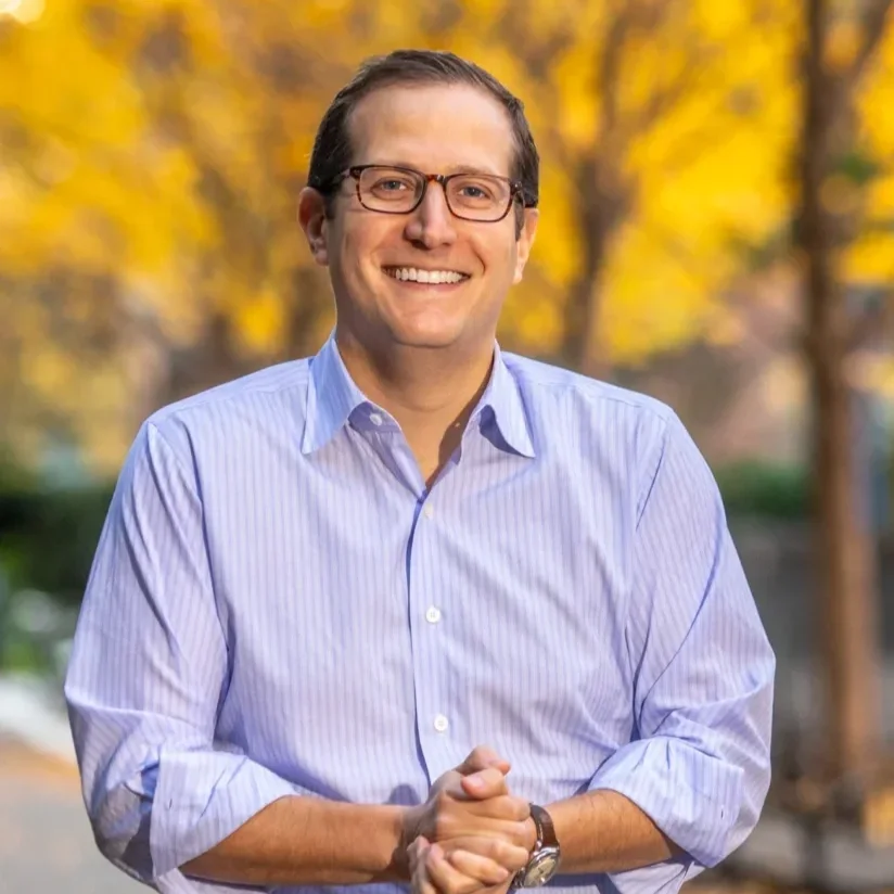 Headshot of Micah Lasher standing outside smiling towards camera with hands clasped