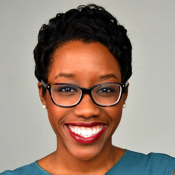 Close-up of a smiling African American woman with short curly black hair, wearing glasses with a black frame, pearl earrings, and a blue top.