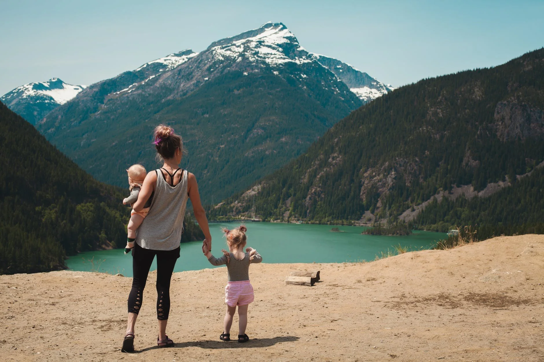 A mom holding a baby and toddler as they all look across a lake at a mountain