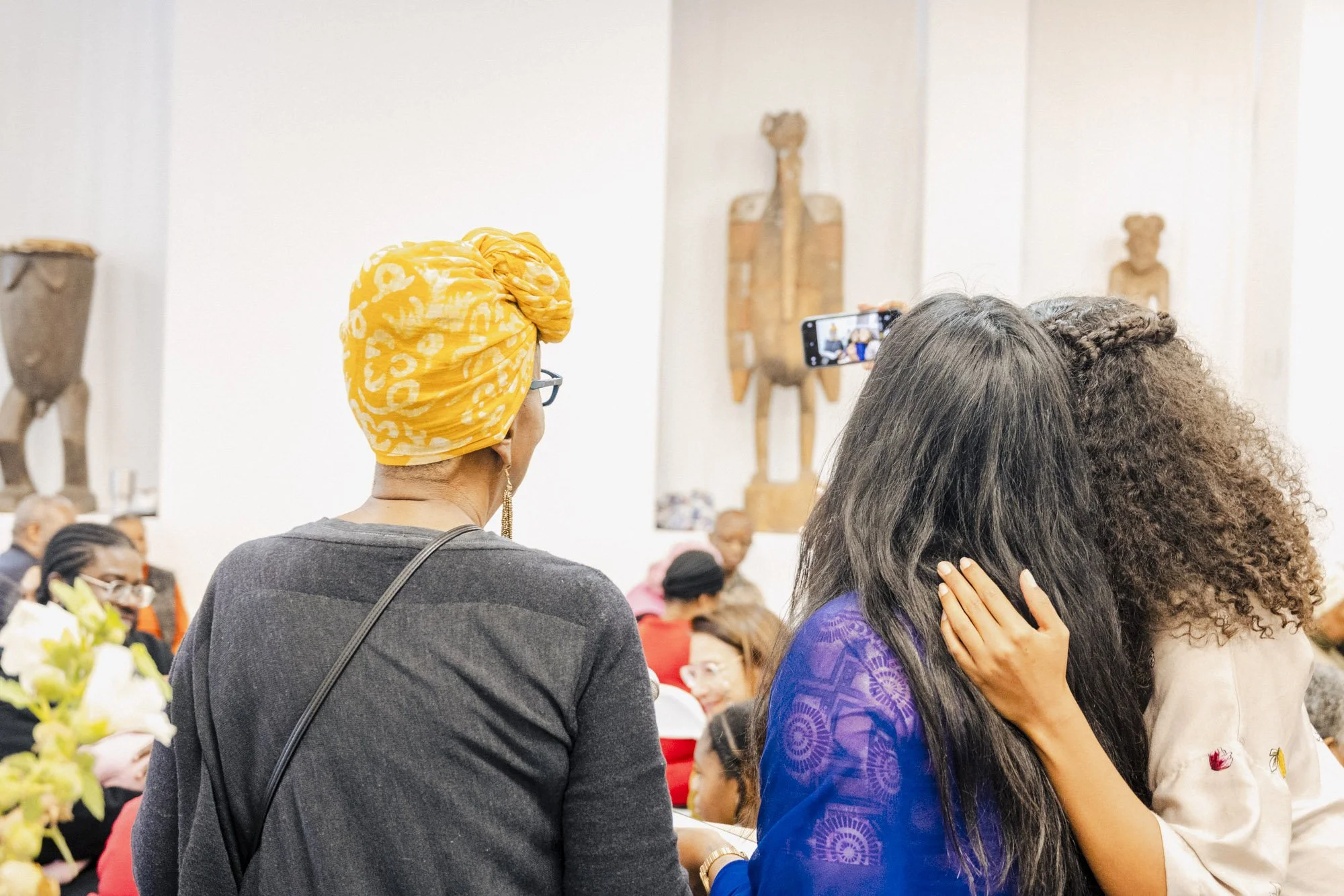 People gathered at an indoor event, with a woman wearing a yellow and white headwrap and glasses, and a woman with long curly hair and a purple dress, sitting and engaged in conversation.