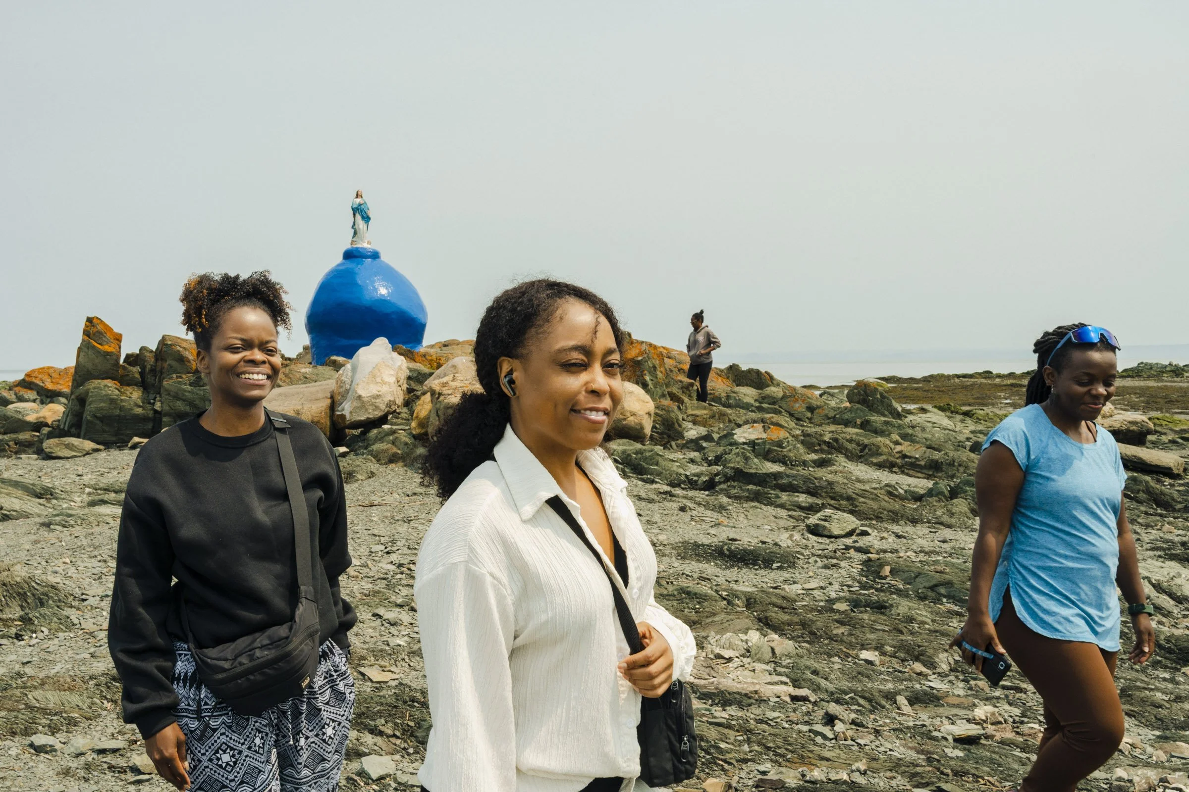 Four women standing on a rocky beach near the ocean, smiling and enjoying a cloudy day. There is a small statue of the Virgin Mary on a blue structure in the background.