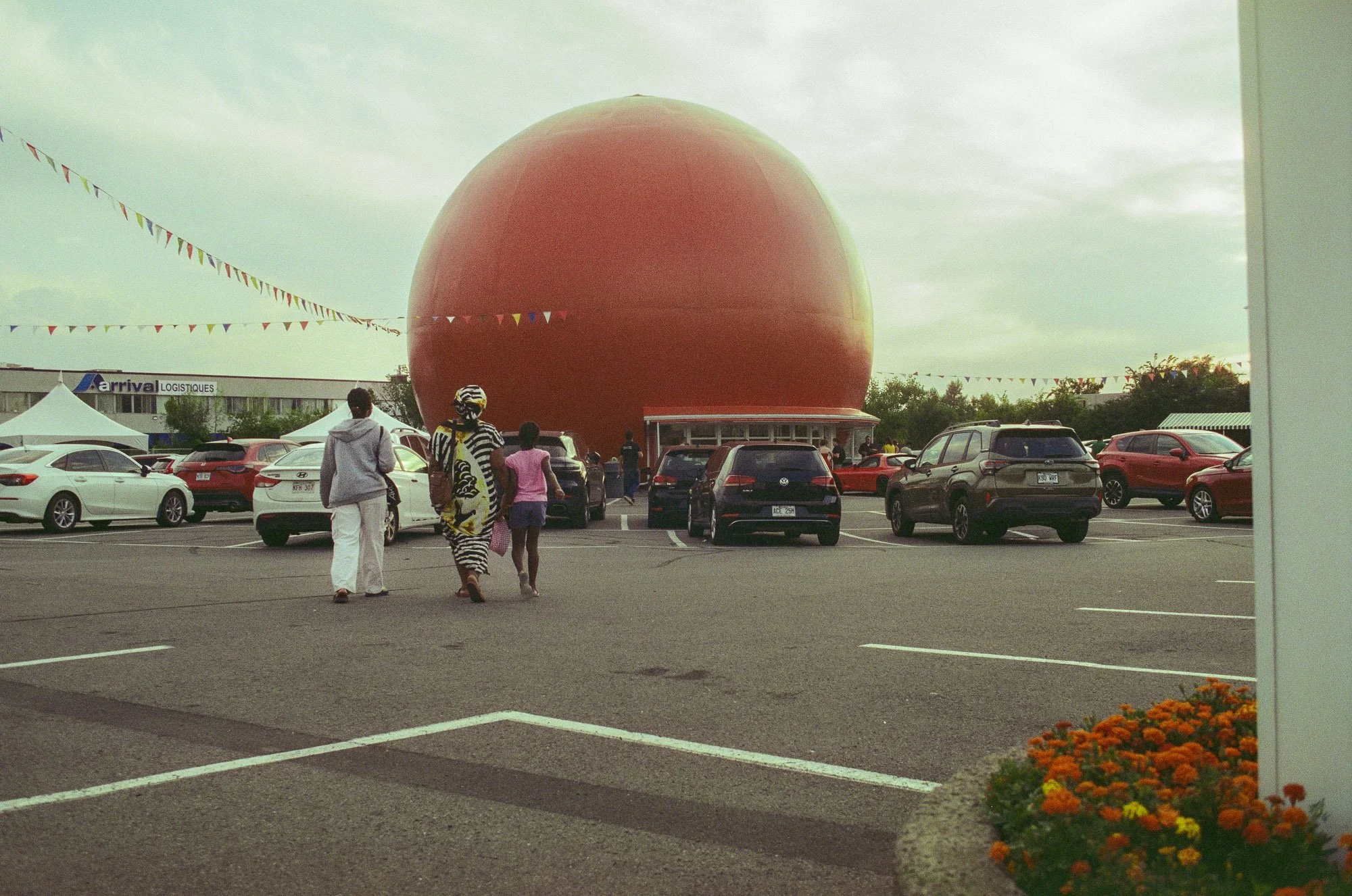 Large orange spherical balloon in a parking lot near tents, with people walking and cars parked, and decorated with colorful bunting flags against a cloudy sky.
