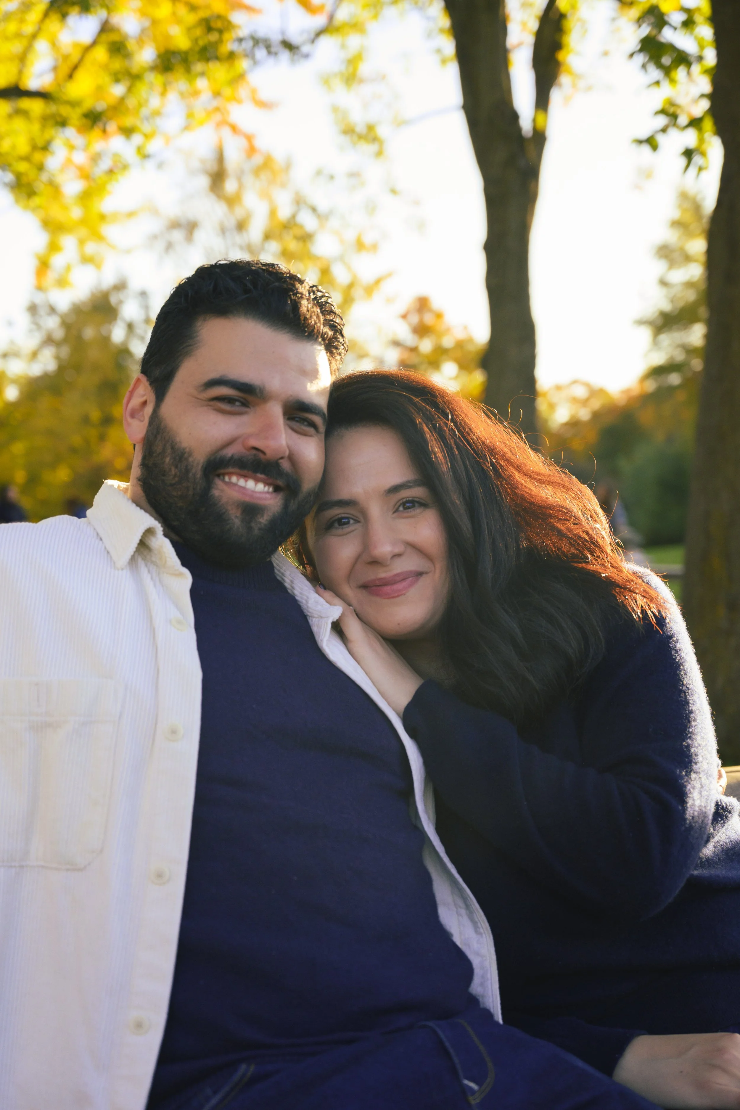 A smiling man and woman embracing outdoors in a park with yellow autumn leaves and trees in the background.