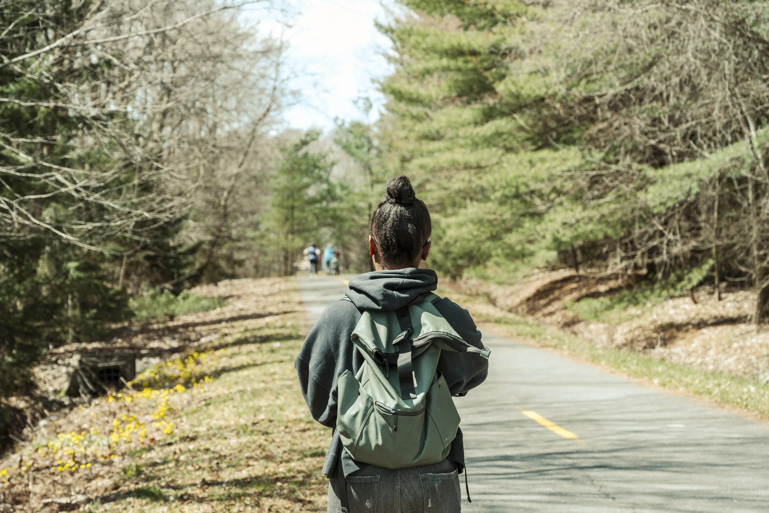 Back view of a woman with a bun hairstyle, wearing a gray hoodie and carrying a backpack, walking on a trail in a forested area with trees and a few other hikers in the background.