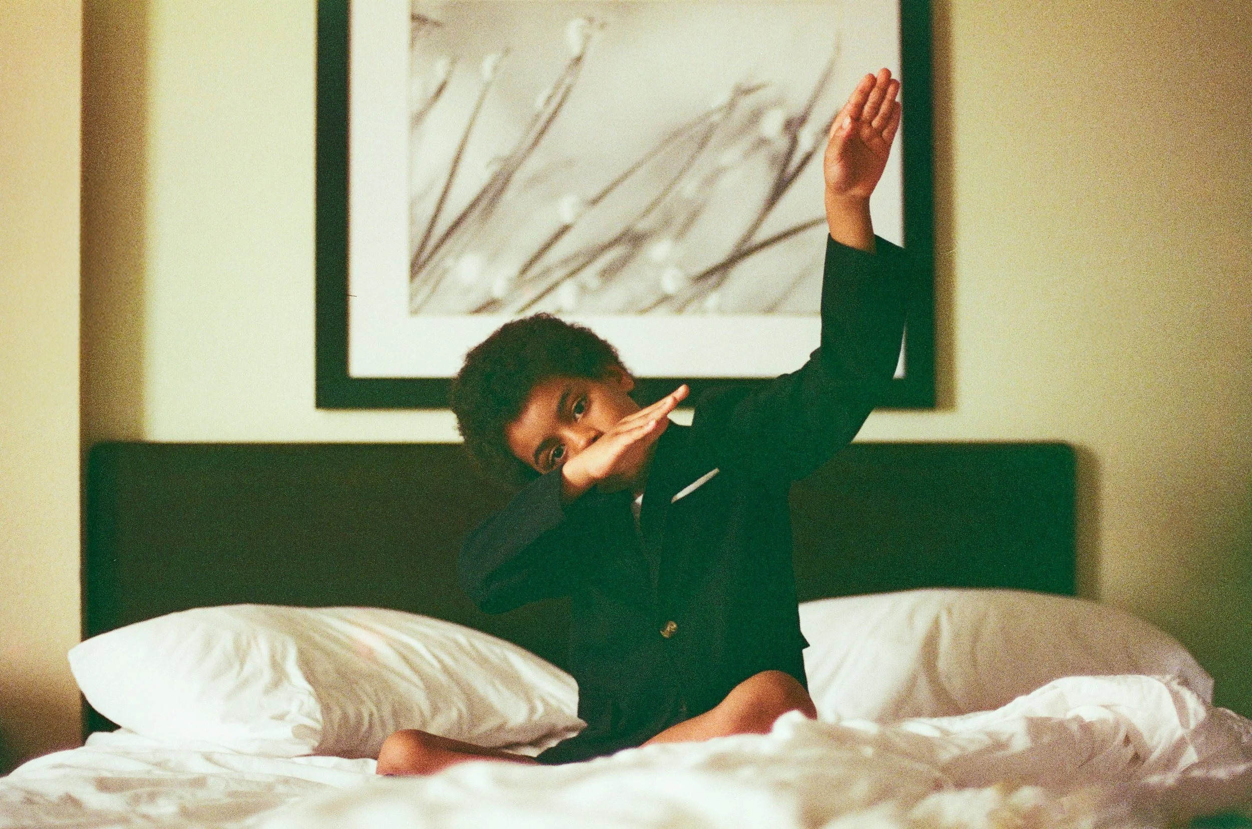 A person with short curly hair sitting on a bed, raising one arm, and using the other arm to cover their face. The person is wearing a black outfit. In the background, there is a framed black-and-white image of tree branches.