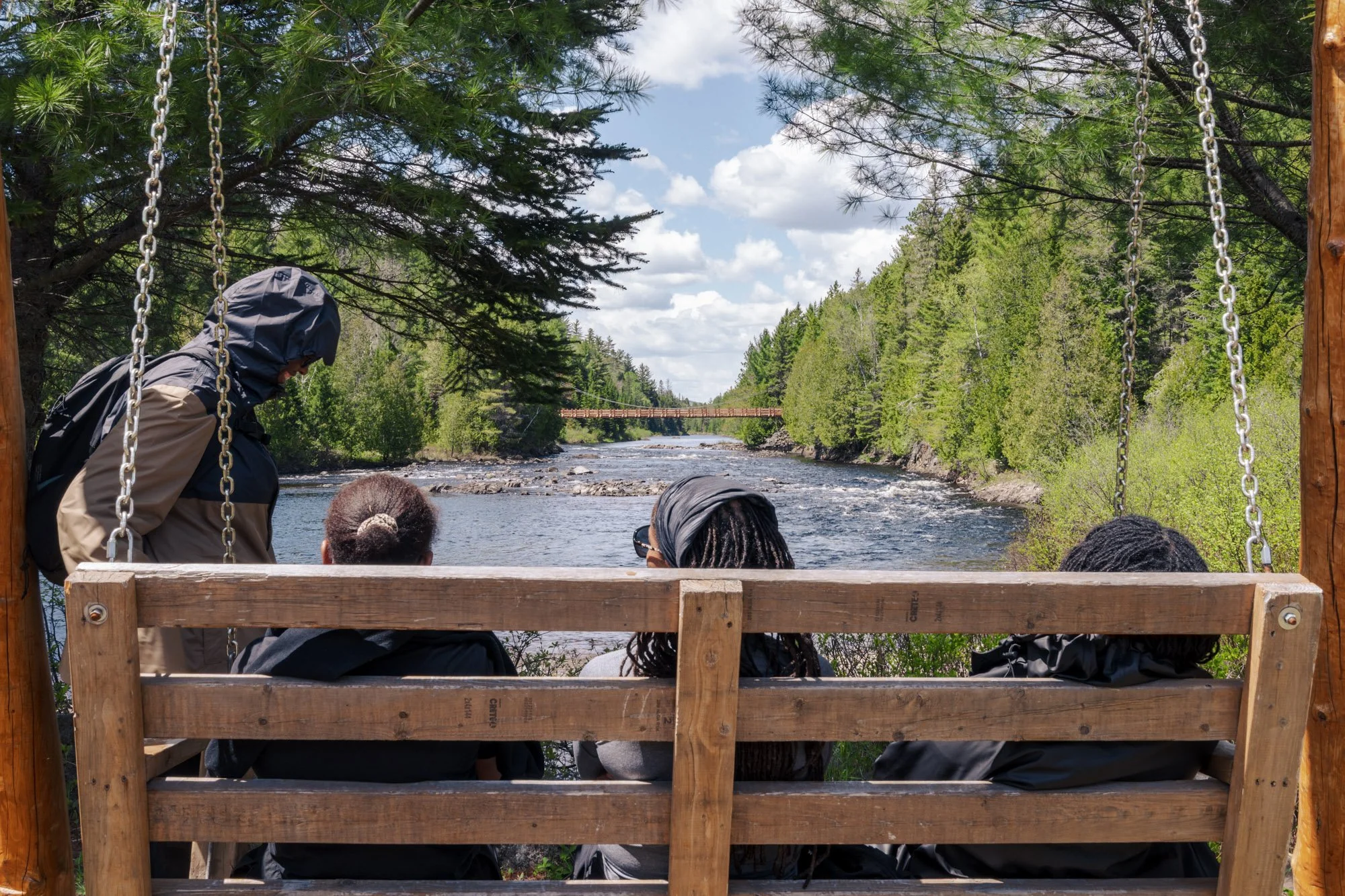 People sitting on a wooden bench with swings overlooking a river, surrounded by green trees under a partly cloudy sky.