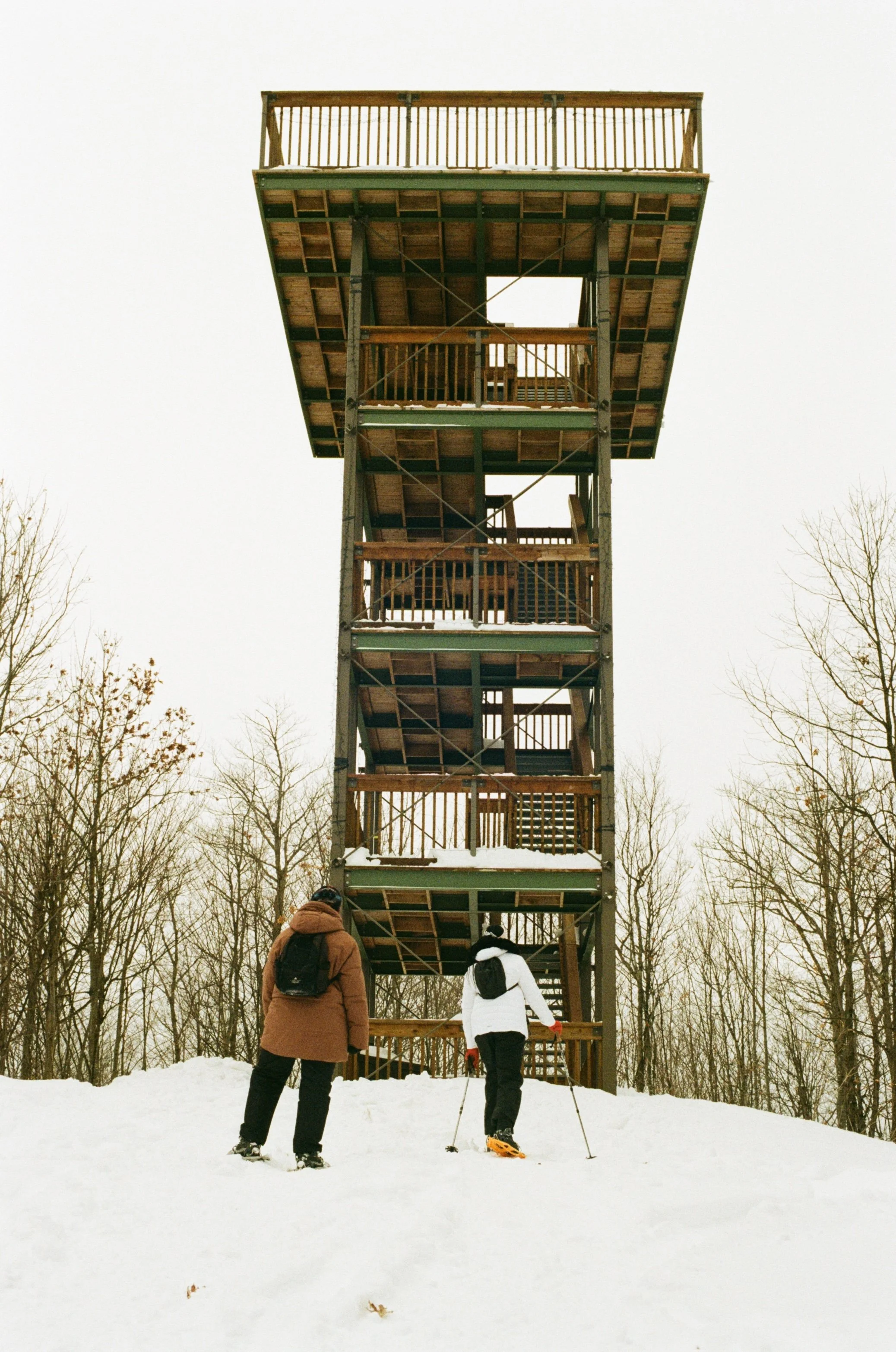 Two people in winter clothing and backpacks preparing to climb a tall wooden observation tower in a snowy park with leafless trees.