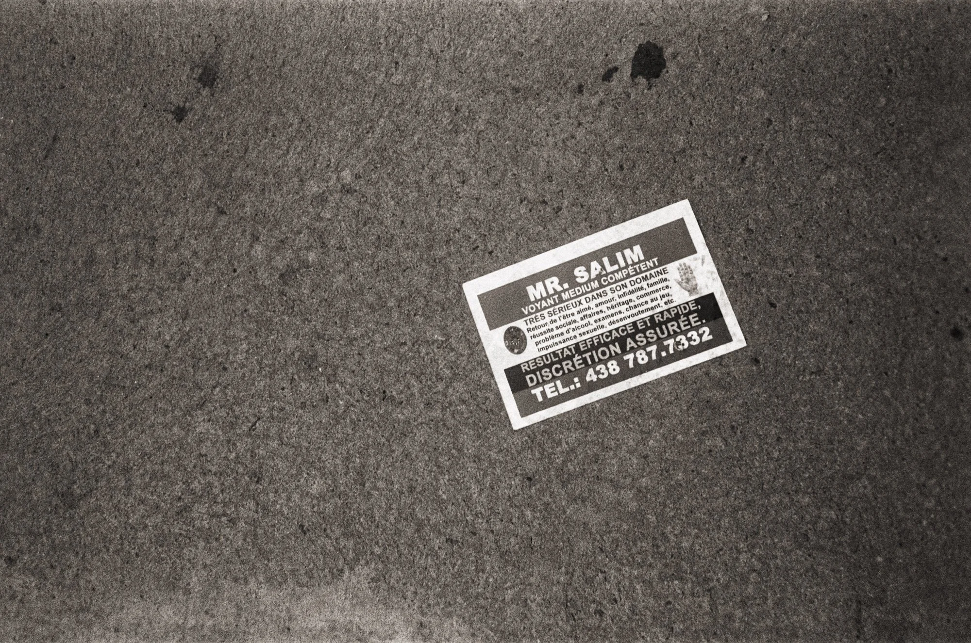 A small sticker on gray pavement has a name, description, and phone number in French, with some black marks on the ground nearby.