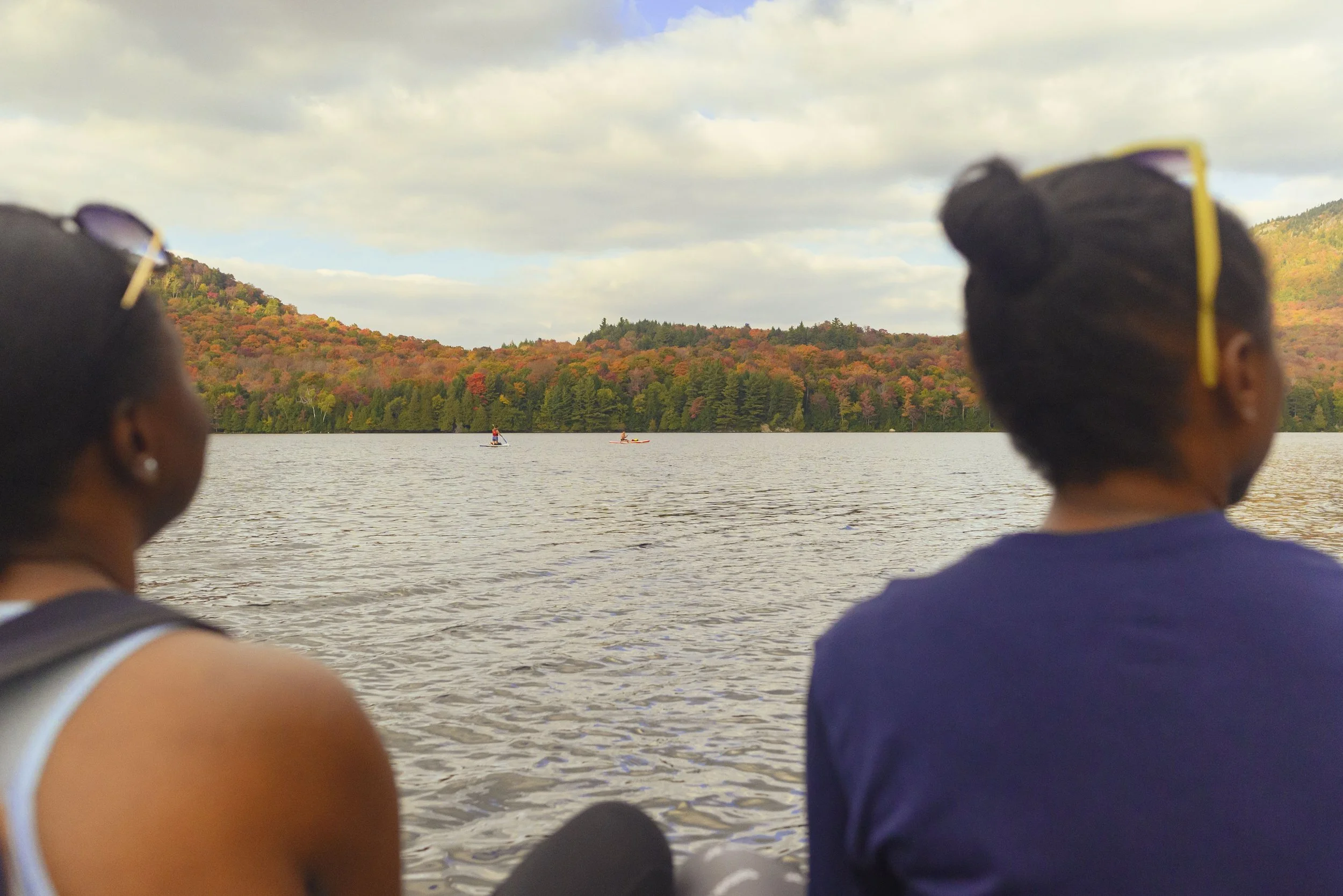 People sitting by a lake with a forested hill showing fall colors in the background, and paddleboarders on the water under a partly cloudy sky.
