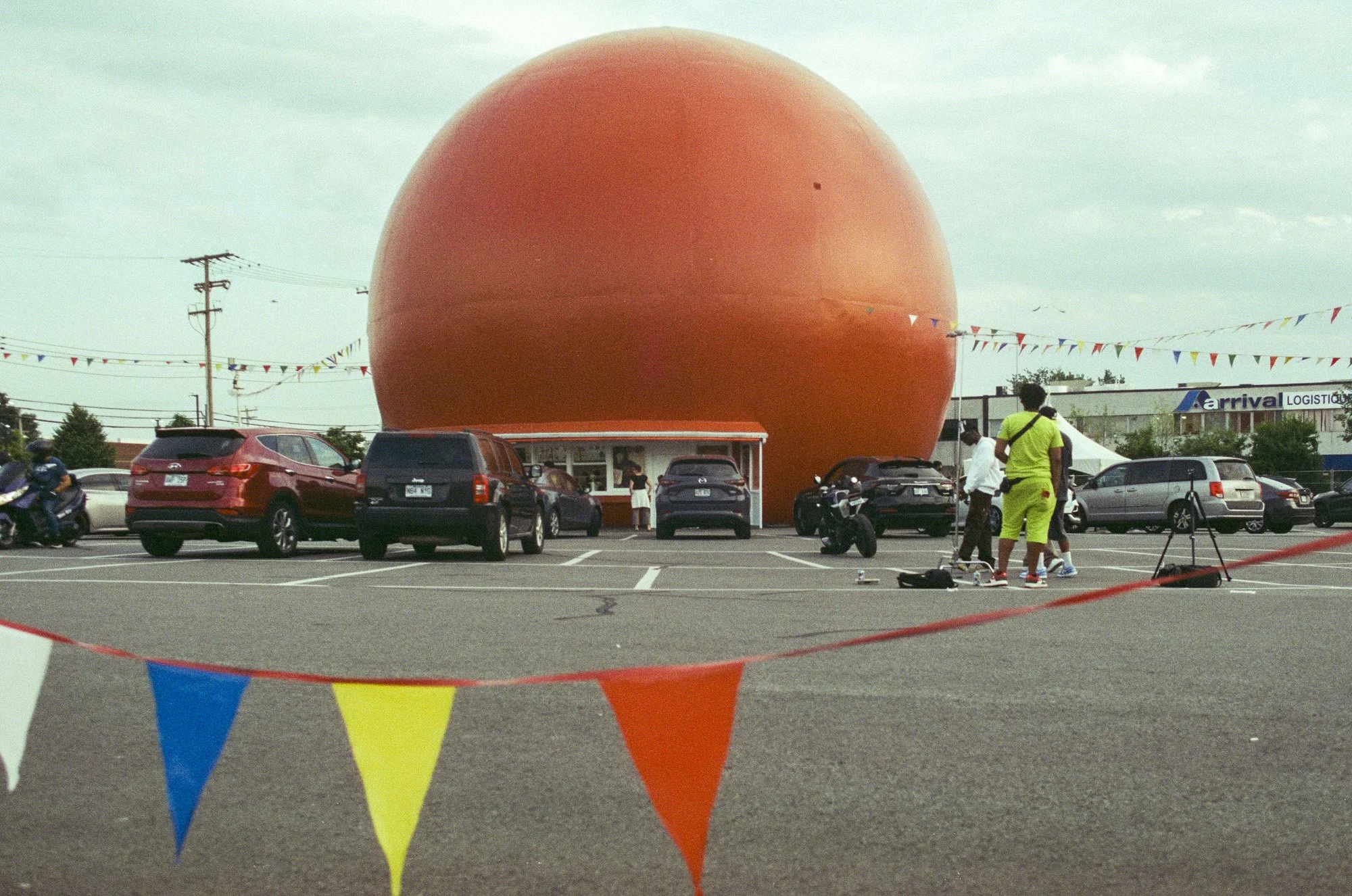 People in a parking lot with a large orange sphere in the background, colorful triangular flags hanging, and a building with an 'arrival LOGISTICS' sign