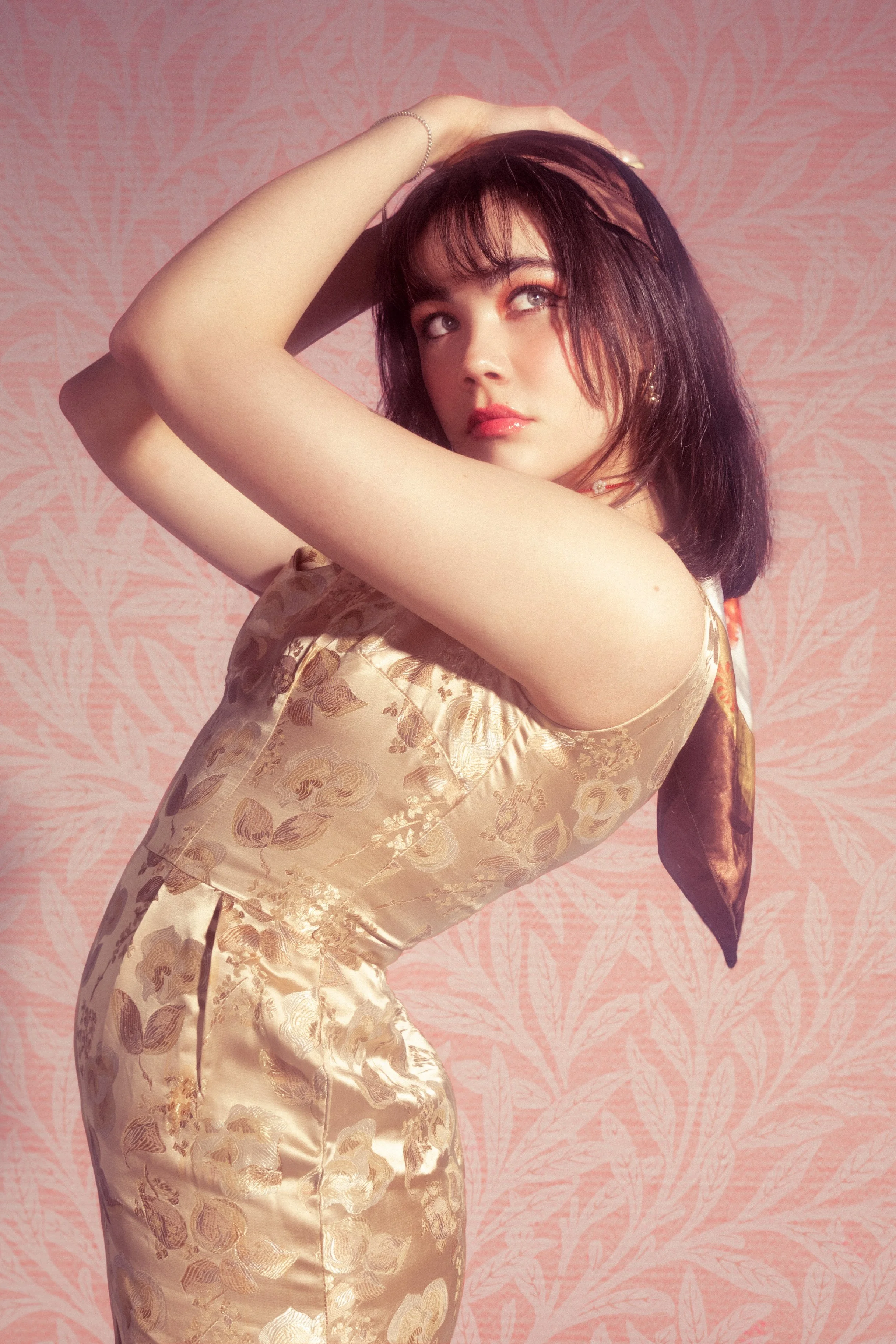 A woman with dark shoulder-length hair, light skin, and wearing a gold satin dress with a floral pattern, poses with one arm above her head against a pink patterned background.
