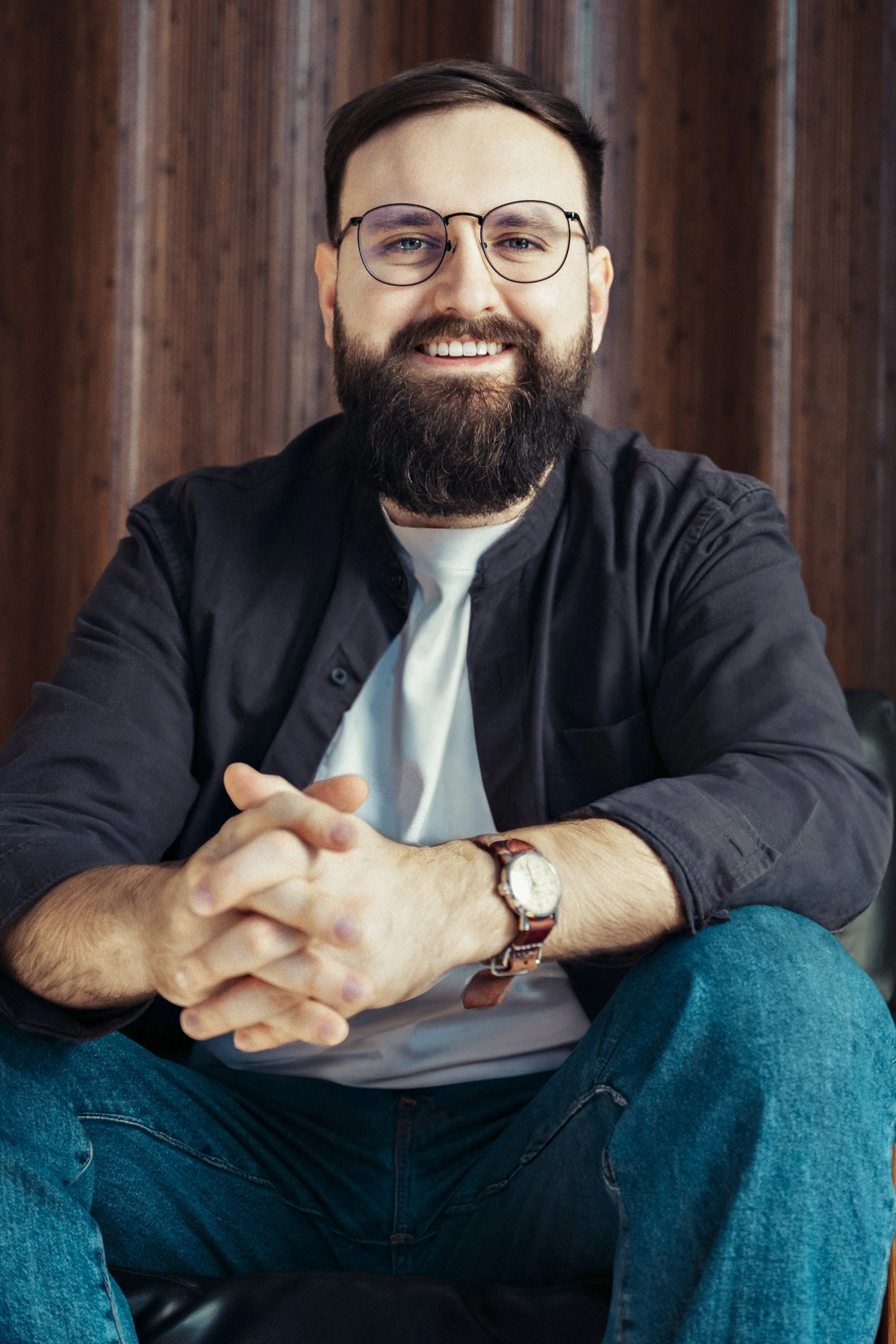 A smiling man with glasses and a beard, wearing a black shirt and a watch, sitting against a wooden wall background.