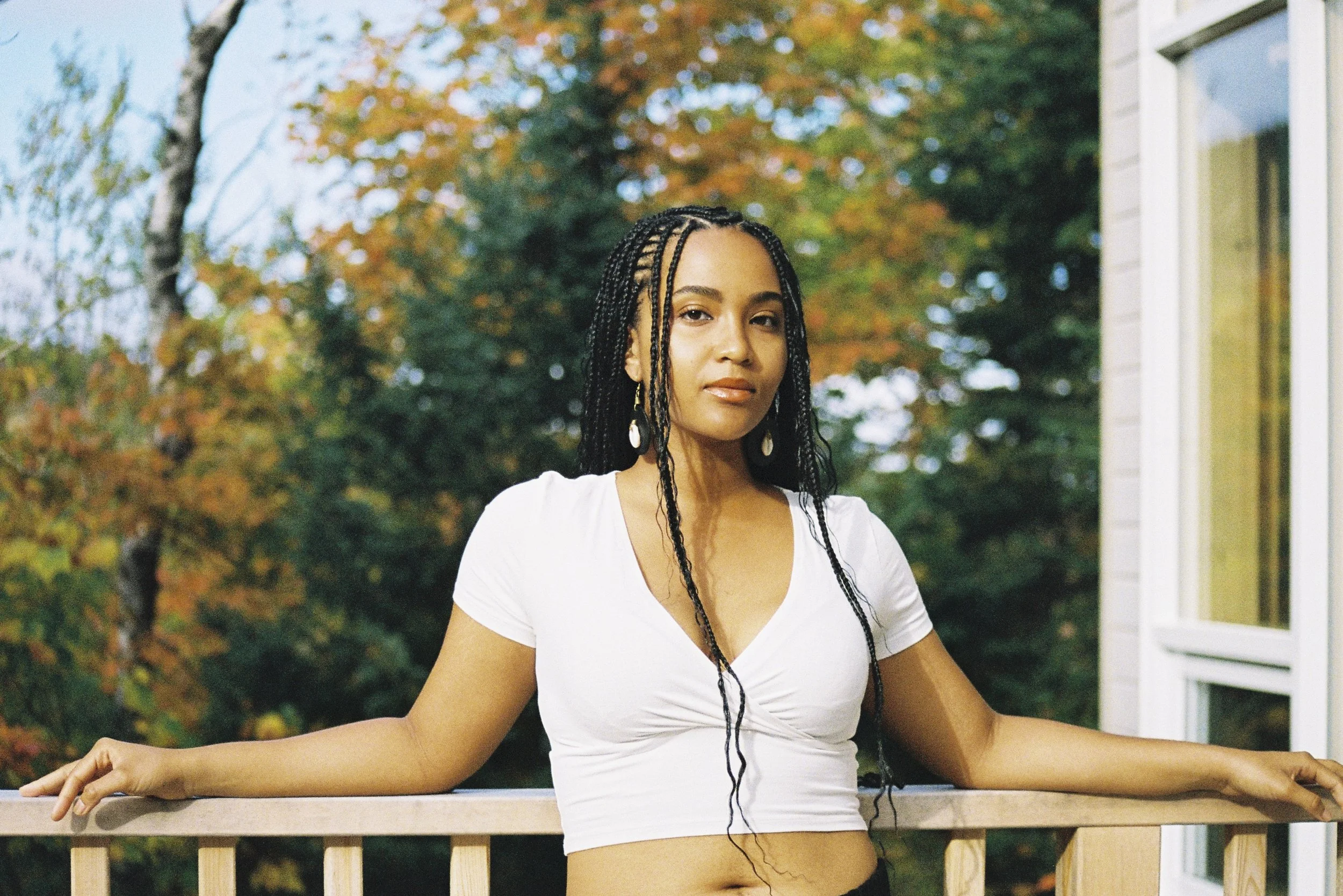 A young woman with long braided hair, wearing a white shirt and earrings, practices yoga outdoors on a wooden deck with autumn trees in the background.