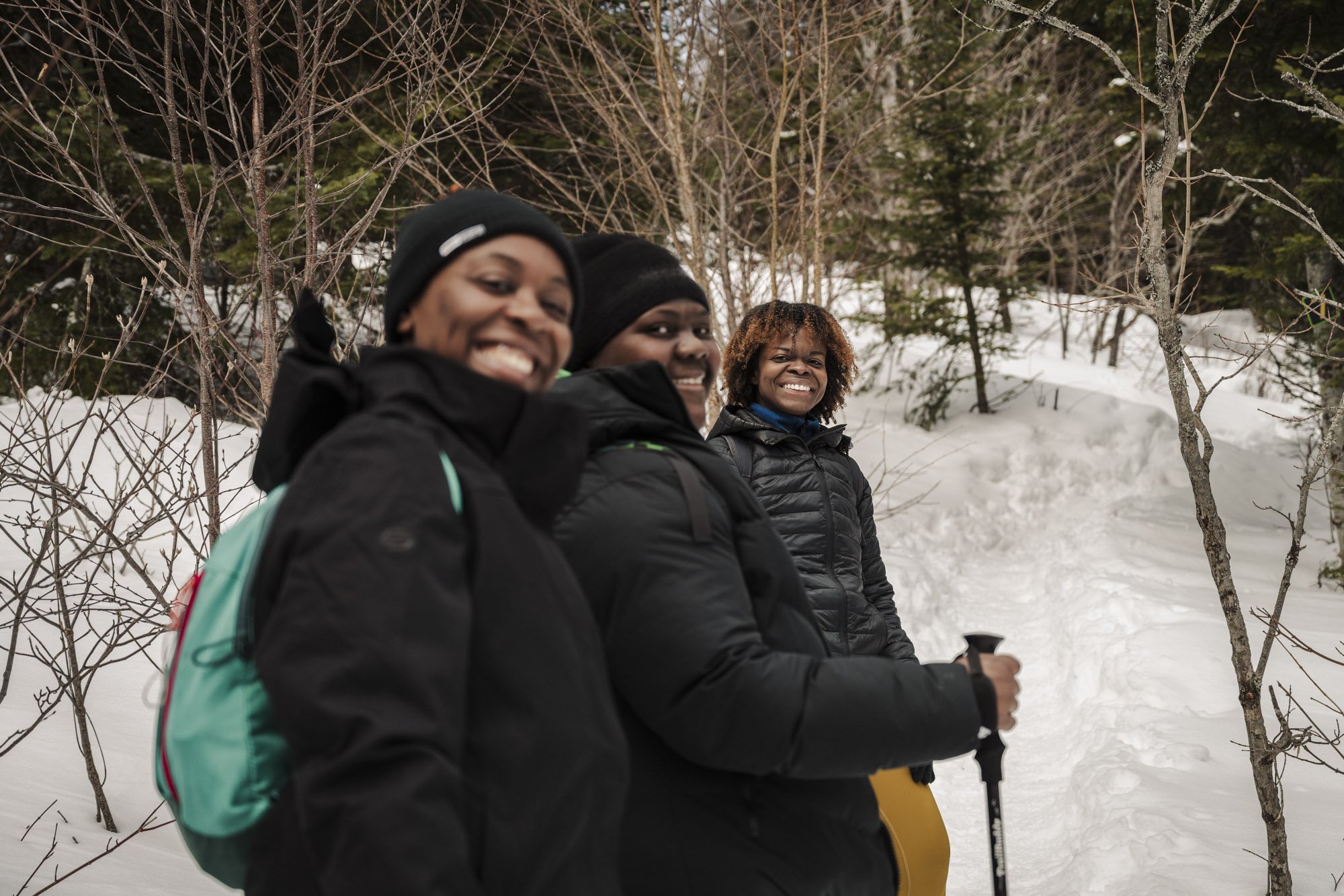 Three women smiling and sitting on a snow-covered ground in a forest during winter, dressed in winter jackets and beanies, with one holding ski poles.