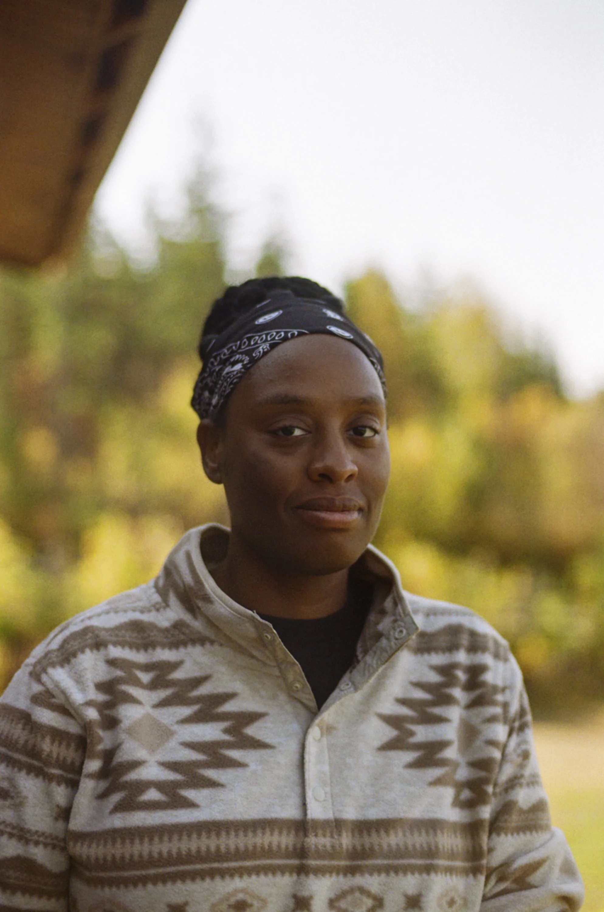 A person with dark hair, wearing a patterned bandana and a beige and brown geometric patterned sweater, outdoors during fall.