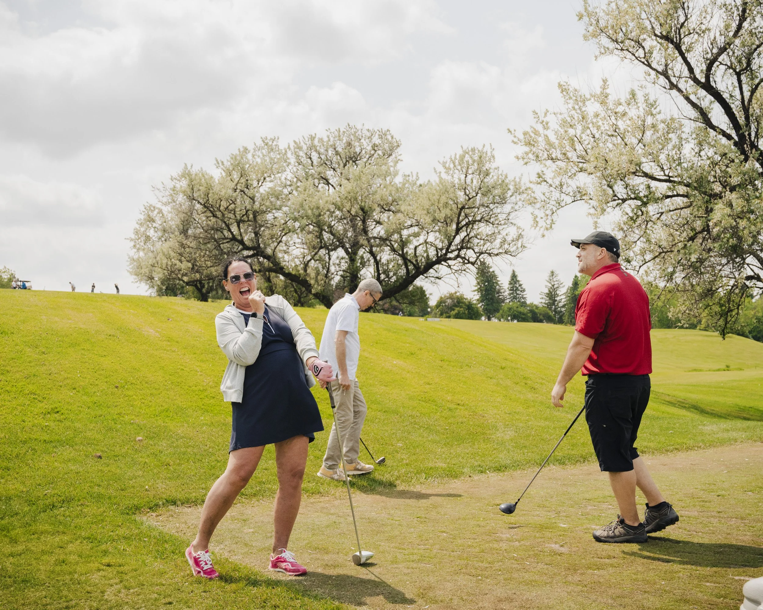 A group of people are playing mini golf on a grassy field with trees in the background. One woman is celebrating with a fist pump, while a man in a red shirt and a cap is holding a golf club and looking at her.